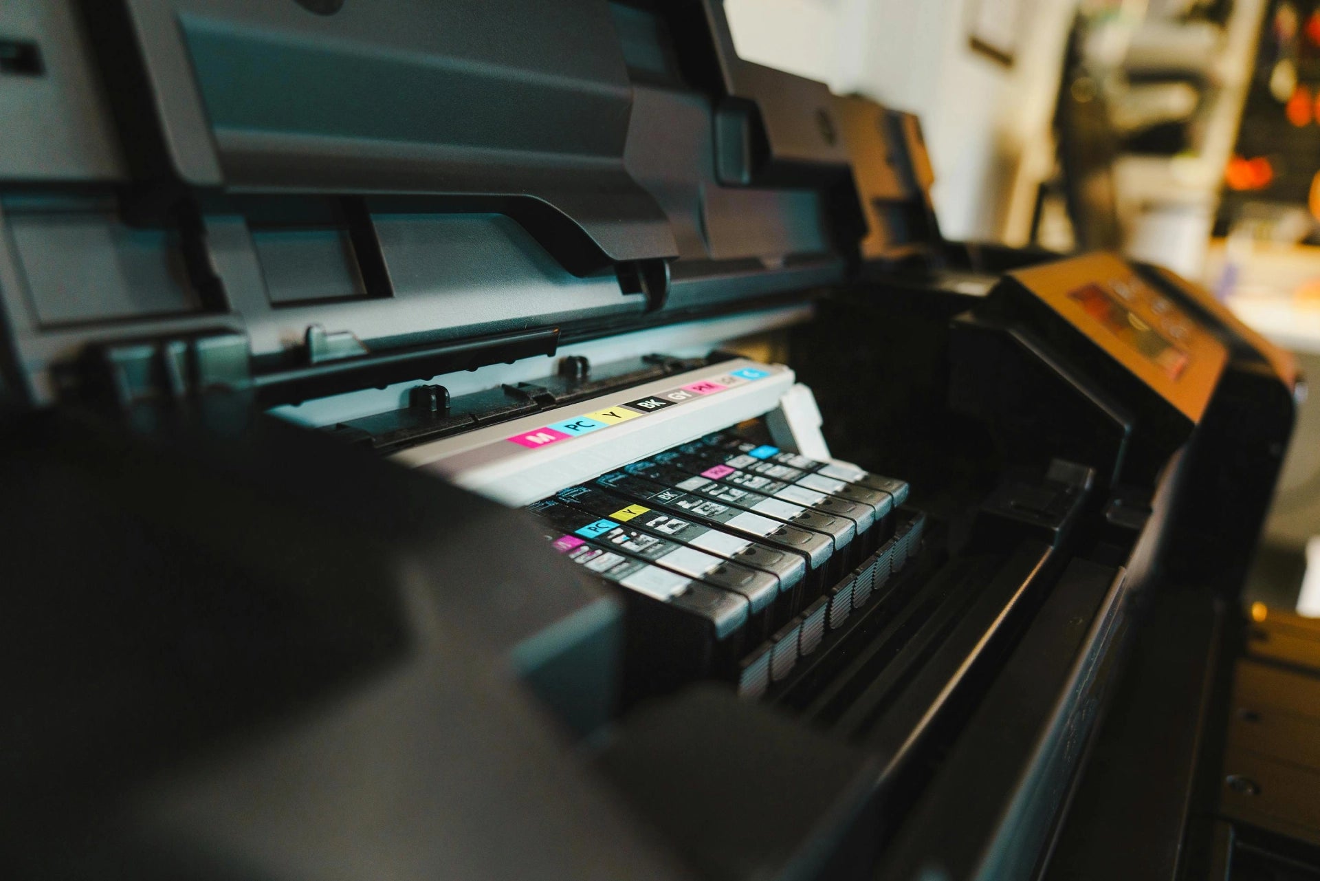 Close-up interior view of a professional wide-format plotter machine, showing a high-capacity inkjet carriage with multiple specialized ink cartridges including Cyan, Magenta, Yellow, Black, and Gray for precision color reproduction.